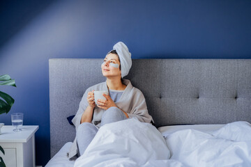 Woman in pajama and bathrobe with eyes patches and towel on her head relaxing and drinking morning...