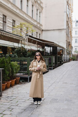 Obraz premium full length of cheerful young woman in trendy sunglasses and trench coat holding paper cup.