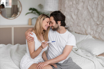 Happy young woman hugging her husband, holding positive pregnancy test on bed at home