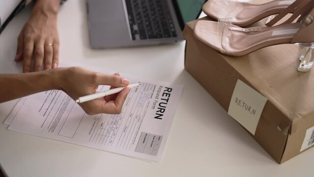 Closeup view of young woman writing return order and sitting at table with laptop in apartment spbd. 4k American African female fills out paper form and wants to return goods back to store, sits at