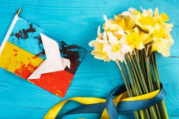 Yellow daffodils with a yellow-blue ribbon on a wooden background and a white paper's dove on ukrainian flag with blood