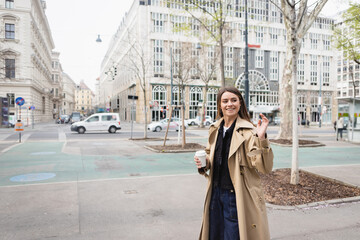 Fototapeta premium happy young woman in stylish trench coat walking with coffee to go and waving hand in european city.