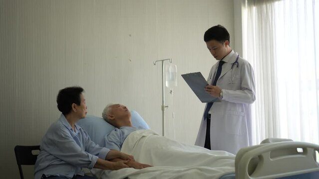Asian young man doctor writing patient details and medical treatment information while checking on old patient