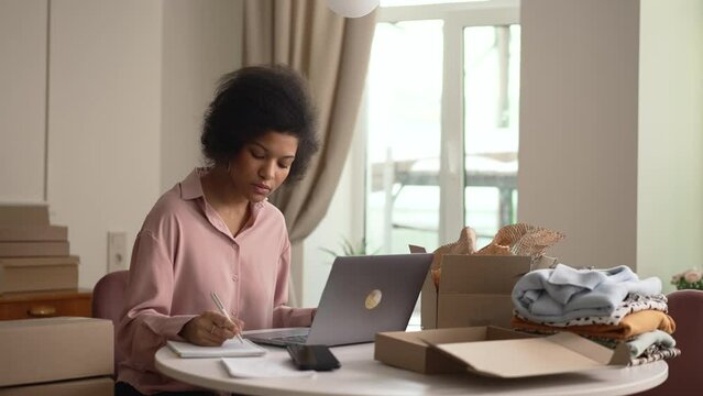 Young woman writing in notebook and preparing clothing orders at table in home office spbd. 4k American African female looks at computer and writes in notebook, sits at desk with packages of clothes