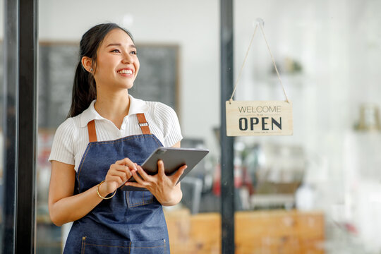 Young Asian businesswoman small business owner standing at cafeteria door entrance pen signboard, A cheerful entrepreneur young waitress in a blue apron near the glass door 