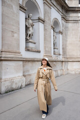 full length of happy young woman in stylish trench coat and baseball cap walking on street of vienna.