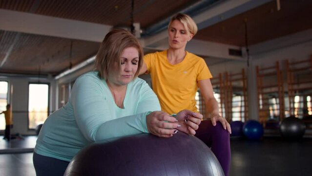 Happy overweight woman exercising with personal trainer on fintess ball in gym