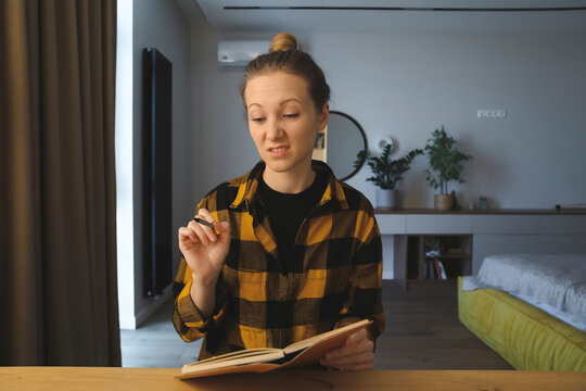 young caucasian woman is sitting at the desk in her bedroom at home, writing notes, making plans in her notebook, holding a pen, showing her dislike with face expression, pulling her face