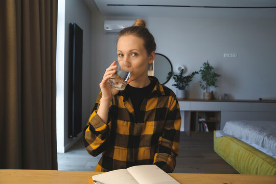 young caucasian woman is sitting at the desk in her bedroom at home, drinking water, looking at camera, wearing checked yellow shirt, hair bun, cozy interior