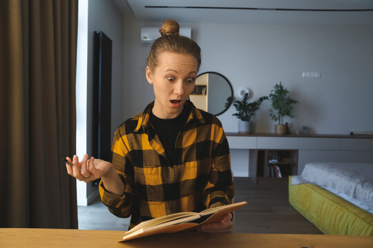 young caucasian surprised woman is sitting at the desk in her bedroom at home, looking at the notebook, amazed with round eyes, holding a pen, wearing checked yellow shirt, hair bun, cozy interior