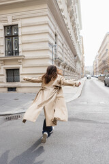 back view of woman in beige trench coat walking with outstretched hands in european city.