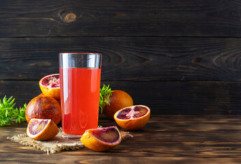 A clear tall glass of red orange juice on a canvas napkin. Red Sicilian oranges and green twigs in the background. Wooden background, space for text