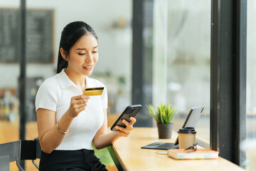 smiling woman paying online, using laptop, holding plastic credit card, sitting on coffee shop, Asian female shopping, making secure internet payment, browsing banking service.
