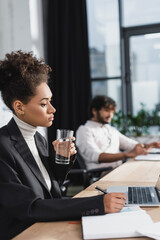 African american businesswoman holding glass of water near notebook and laptop in office.