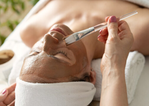 The face of a white woman undergoing cosmetic procedures. Brown clay face mask. Facial care. Applying facial mask at woman face at beauty salon