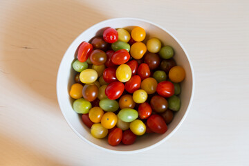 Selective focus high angle side lit flat lay view of red, yellow and green cherry tomatoes in white bowl on pale wooden background
