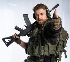 the airsoft player puts his rifle on his shoulder and aims at us with his fingers. a man in an outfit, in headphones, a bulletproof vest, with a backpack on a white background. - obrazy, fototapety, plakaty