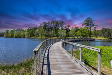 A long winding brown wooden bridge over a rippling blue lake surrounded by lush green trees, grass...