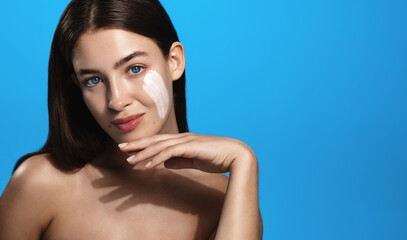 Beautiful girl model with clay mask, lotion or skin care facial product on cheek, using cosmetic mask, clay for nourished, glowing skin effect, blue background. High quality