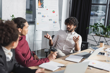 Indian businessman talking to african american colleagues near devices and flip chart in office.