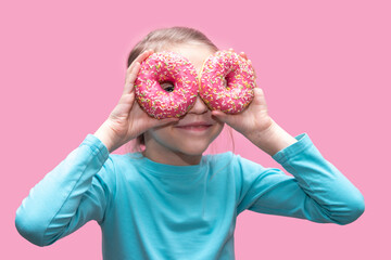 A cute funny girl in a blue t-shirt holds bright pink donuts near her eyes like glasses and looks at you on a pink background. Cheerful childhood concept. Sweet food for kids concept