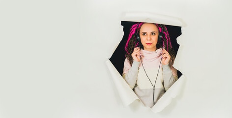 Young woman in headphones in hole of white background. Beautiful female with curly hair listening to music in studio.