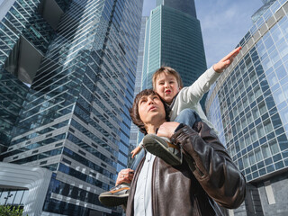 Fototapeta premium Laughing boy plays like airplane on father's shoulders. Dad and son looks on glass walls of buildings. Future and modern technologies, life balance and family life in well keeps districts.