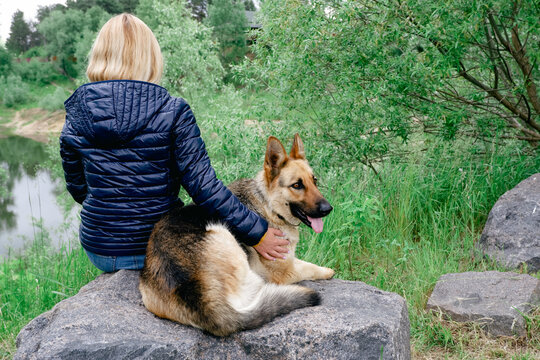 A young woman with a German Shepherd sits on a large granite stone by the lake on a cloudy spring day. Young woman relaxing with her friend dog at the coast sitting on a stone promenade looking out
