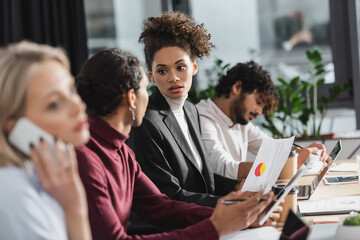 African american businesswoman holding paper near colleague with digital tablet in office.