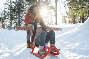 Joyful African American man and woman in love having fun on winter day sledding in local mountains