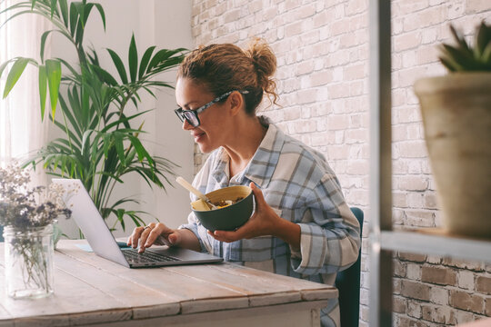 Woman Working At Home Office Table Eating Healthy Food In A Bowl. Independent Female People Lifestyle, Alternative Job Occupation Using Laptop Computer And Online Opportunity. Modern Worker Business