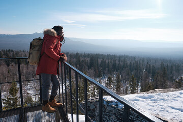 Fototapeta na wymiar Young African American man wearing backpack standing relaxed at observation deck on mountain top on winter day