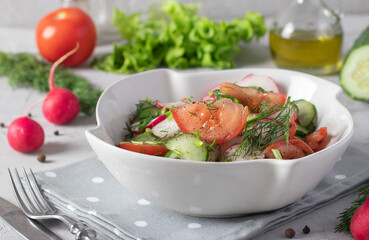 Salad with cucumbers, tomatoes and radishes dressed with olive oil in a white salad bowl on a gray background