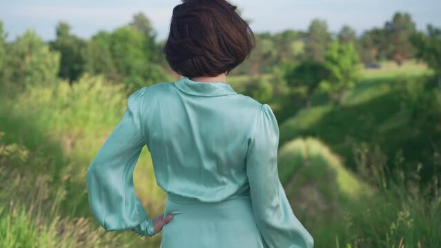 Back view confident slim woman standing outdoors admiring beauty of nature on summer spring day. Relaxed Caucasian elegant lady enjoying leisure in park outdoors on sunny day. Slow motion
