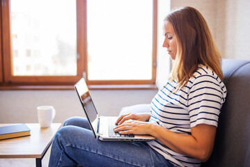 Young woman with a laptop and a cup of coffee on the table at home. Woman looking at laptop screen, reading good news in message, watching video, chatting in social network or shopping online