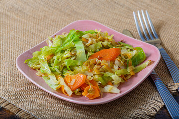 Carrots fried with cabbage, herbs and onions on a pink square plate, fork and knife on a burlap tablecloth on the table. Copy space. Vegetarian food.