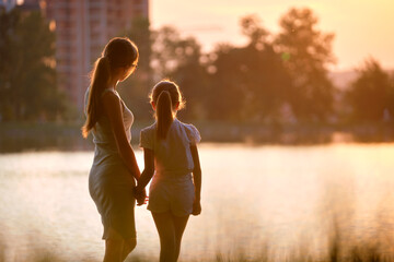 Happy mother and daughter standing together holding hands enjoying time in summer park in evening....