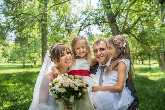 The groom in a suit and the bride in a long white dress with children in summer dresses in a spring garden on green grass. Happy family on a warm sunny day among the trees.