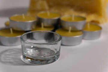 A pile of beeswax tealight candles are displayed with raw beeswax in the background. A clear glass holder is in the front.