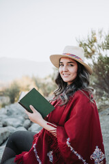 Young traditional hispanic woman using a hat and a red poncho, sitting in a natural space and reading a book.