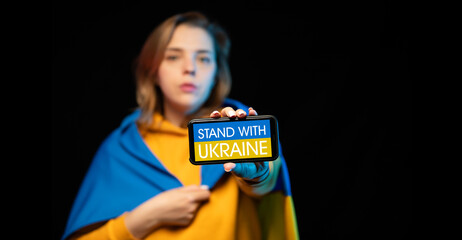Beautiful Ukrainian girl with a state flag and smartphone in hands with a call STAND WITH UKRAINE. The actual concept of anti-war support. Russian-Ukrainian war. Prays for Ukraine. STOP WAR