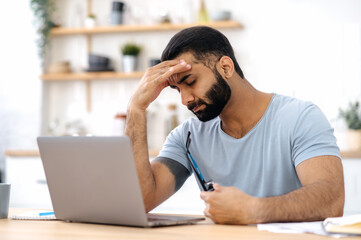 Exhausted indian or arabian young adult man, freelancer, designer, in casual wear, sit in the...
