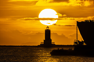 Lighthouse with sun rising above and vessel to right.