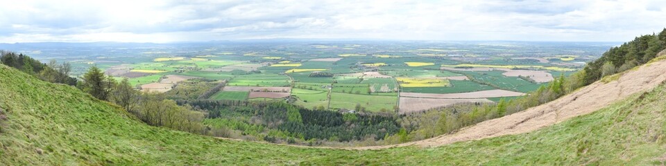 Large panoramic view taken from the top of The Wrekin hill 407 meters above sea level in Shropshire, England, UK. Green, brown and yellow fields fill the countryside with Welsh hills in the background