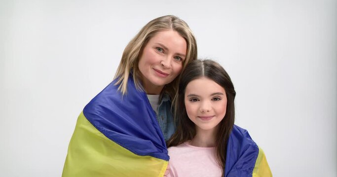Pleasant woman embracing her adorable daughter while standing together in studio and holding Ukrainian flag on shoulders. Family unity, support and freedom concept.