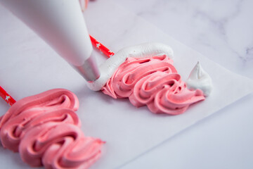 On light parchment there are pink meringue caps on sticks, the cook makes a pompom and a frill from a pastry bag of white meringue. Photography in a light key