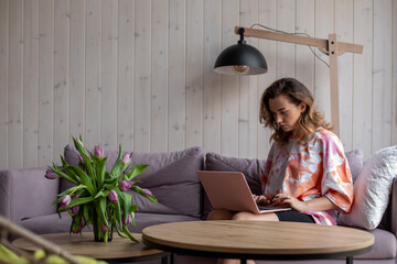 Portrait of woman typing on keyboard of laptop and sitting on soft sofa in casual clothes near table with vase of fading tulips. Living room in elegant design with aesthetic decor, free copy space