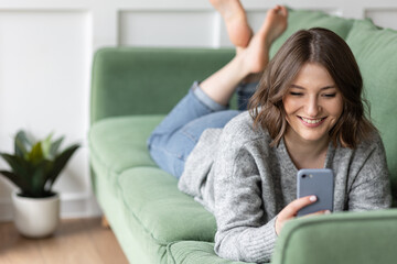 Obraz premium Young beautiful smiling woman using smart phone lying on the couch. Concept of freelance job, home office. Female using technology for learning, shopping, e-commerce, communication, social media