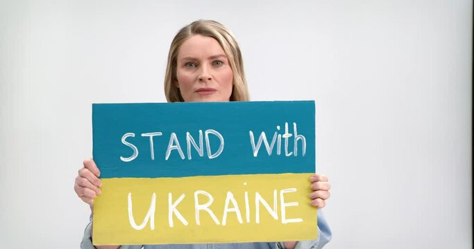 Caucasian woman with blond hair standing in studio with white background and holding banner with lettering Stand with Ukraine. Stop war concept. Pray for Ukraine.