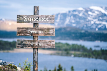 success dreams fortune text quote written on wooden signpost outdoors in nature with lake and mountain scenery in the background. Moody feeling.